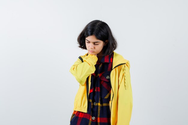 Young girl putting hand on neck in checked shirt and yellow jacket and looking tired. front view.