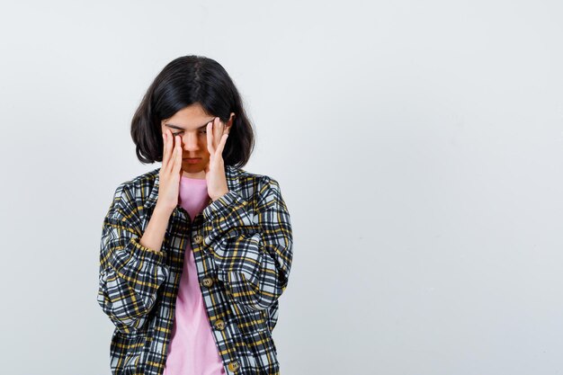 Young girl putting hands on face, rubbing temples while closing eyes in checked shirt and pink t-shirt and looking tired