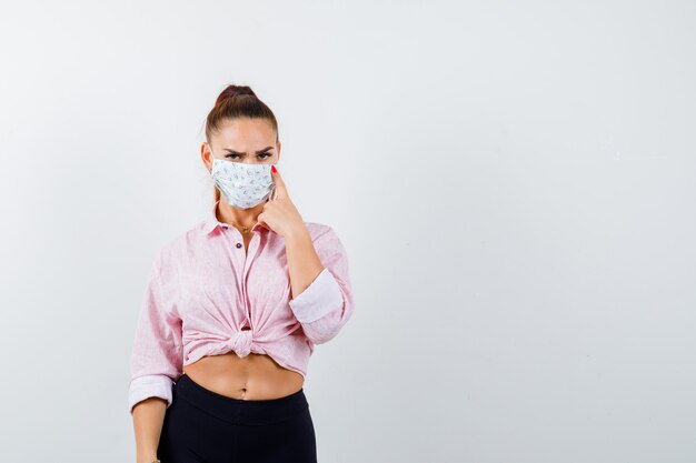 Young girl putting index finger on cheek in pink blouse, black pants, mask and looking confident , front view.