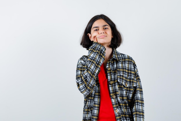 Young girl putting index finger on cheek in red t-shirt and checked shirt and looking cute. front view.