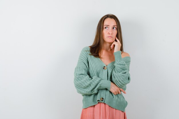 Young girl putting index finger on cheek, thinking about something in knitwear, skirt and looking pensive. front view.