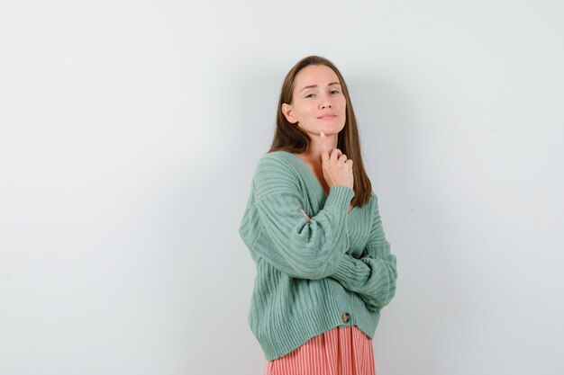 Young girl putting index finger on chin, holding hand on arm in knitwear, skirt and looking alluring , front view.