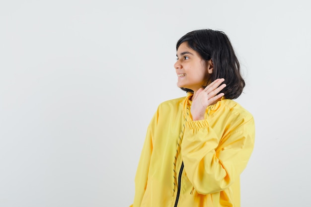 Young girl raising hand as greeting someone in yellow bomber jacket and looking happy.