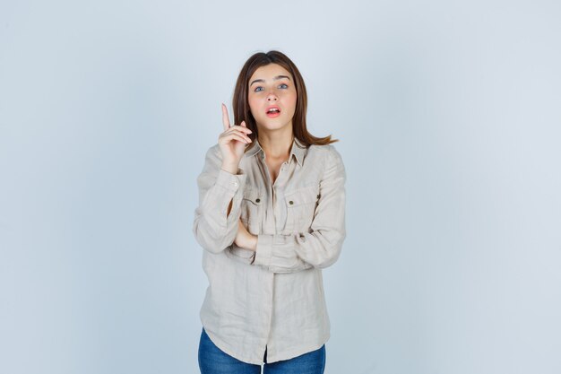 Young girl raising index finger in eureka gesture in beige shirt, jeans and looking sensible , front view.