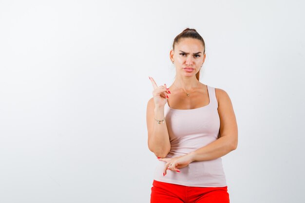 Young girl raising index finger in eureka gesture in beige top and red pants and looking serious , front view.