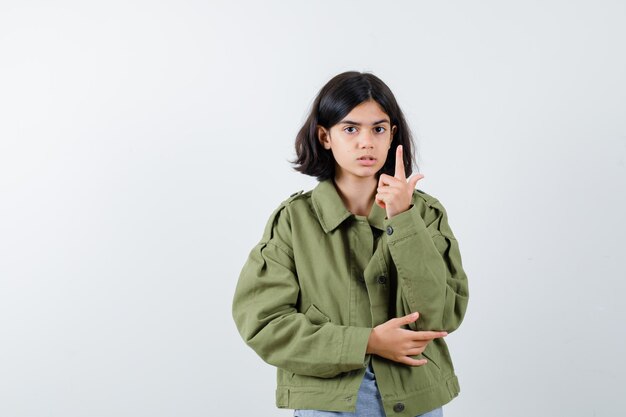 Молодая девушка поднимает указательный палец в жесте эврики в сером свитере, куртке цвета хаки, джинсовых брюках и выглядит мило. передний план.