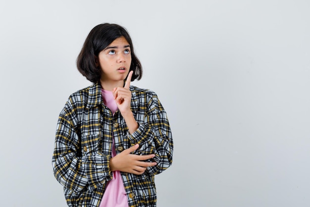 Free photo young girl raising index finger in eureka gesture while holding hand on elbow in checked shirt and pink t-shirt and looking pensive. front view.