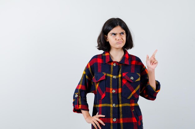 Young girl raising index finger in eureka gesture while holding hand on waist in checked shirt and looking pensive. front view.