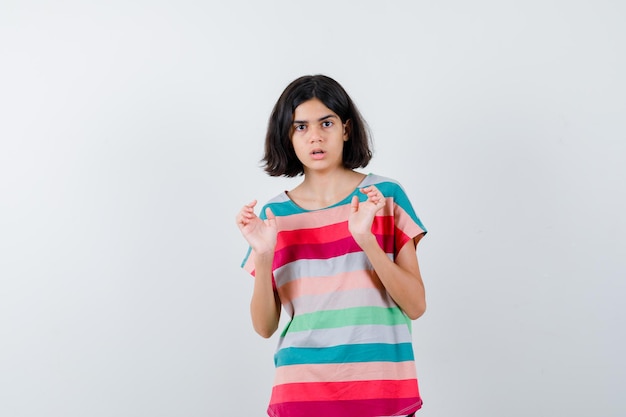 Young girl raising palms in surrender gesture in colorful striped t-shirt and looking surprised. front view.