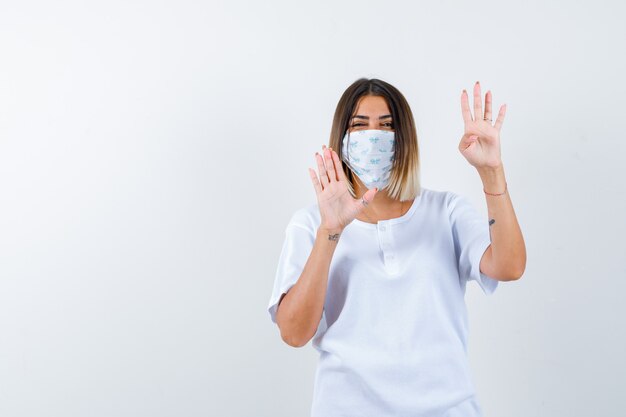 Young girl raising palms in surrender gesture in white t-shirt and mask and looking confident , front view.