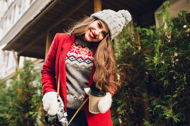 Young girl in red coat walking on street with coffee to go.  She wears white gloves, smiling .