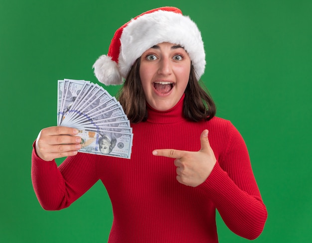 Free photo young girl in red sweater and santa hat holding cash pointing with index finger at money happy and surprised standing over green wall