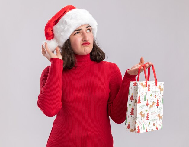 Молодая девушка в красном свитере и шляпе санта-клауса держит красочный бумажный пакет с рождественскими подарками, глядя вверх со скептическим выражением лица, стоя на белом фоне