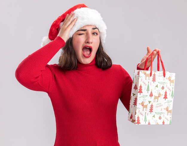 Young girl in red sweater and santa hat holding colorful paper bag with christmas gifts  surprised standing over white wall