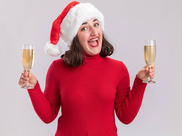 Free photo young girl in red sweater and santa hat holding two glasses of champagne looking at camera happy and cheerful smiling standing over white background