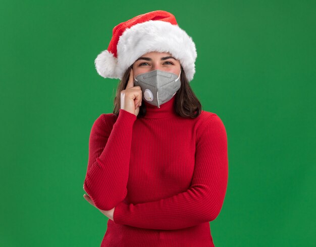 Young girl in red sweater and santa hat wearing facial protective mask  with pensive expression thinking standing over green wall