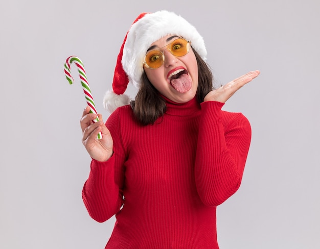 Giovane ragazza in maglione rosso e cappello da babbo natale con gli occhiali tenendo il bastoncino di zucchero felice e allegro che attacca fuori la lingua in piedi su sfondo bianco