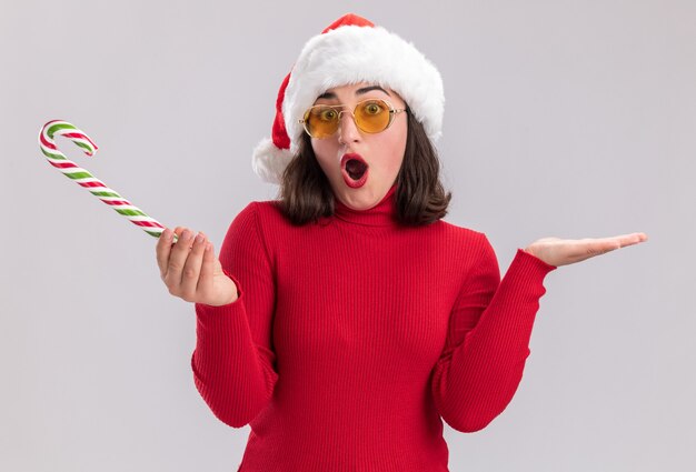 Free photo young girl in red sweater and santa hat wearing glasses holding candy cane looking at camera surprised and confused with arms raised standing over white background