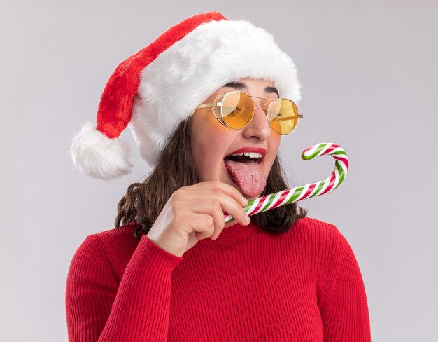 Young girl in red sweater and santa hat wearing glasses holding candy cane trying to taste it happy and joyful standing over white background