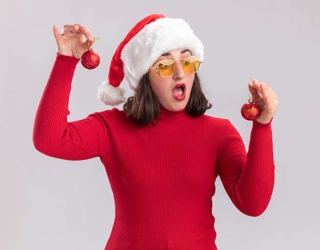 Giovane ragazza in maglione rosso e cappello da babbo natale con gli occhiali che tengono le palle di natale guardandoli confusi cercando di fare la scelta in piedi sul muro bianco