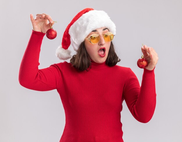 Young girl in red sweater and santa hat wearing glasses holding christmas balls looking at them confused trying to make choice standing over white wall