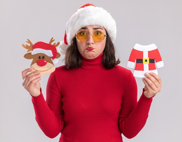 Young girl in red sweater and santa hat wearing glasses holding christmas toys  confused standing over white wall