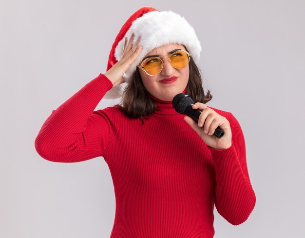 빨간 스웨터와 마이크를 들고 안경을 쓰고 산타 모자에 어린 소녀는 흰 벽 위에 서서 혼란스럽고 불쾌하게 서 있습니다.