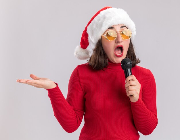 Молодая девушка в красном свитере и шляпе санта-клауса в очках с микрофоном, глядя в камеру, удивлена и поражена рукой, стоящей на белом фоне