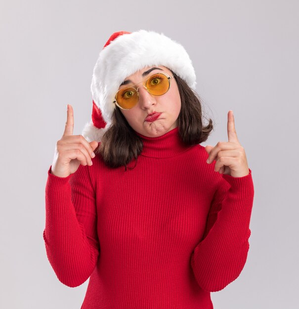 Young girl in red sweater and santa hat wearing glasses looking at camera with sad expression showing index fingers pursing lips standing over white background