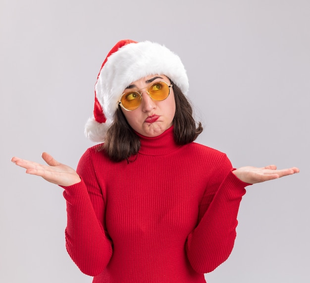 Young girl in red sweater and santa hat wearing glasses looking confused spreading arms to the sides standing over white background