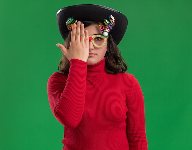 재미있는 안경과 손으로 한쪽 눈을 덮고 검은 모자를 쓰고 빨간 스웨터에 어린 소녀