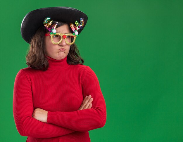 재미 있은 안경과 검은 모자를 쓰고 빨간 스웨터에 어린 소녀가 팔에 불쾌감을 느끼고 녹색 벽 위에 서있는 뺨을 불고 넘어