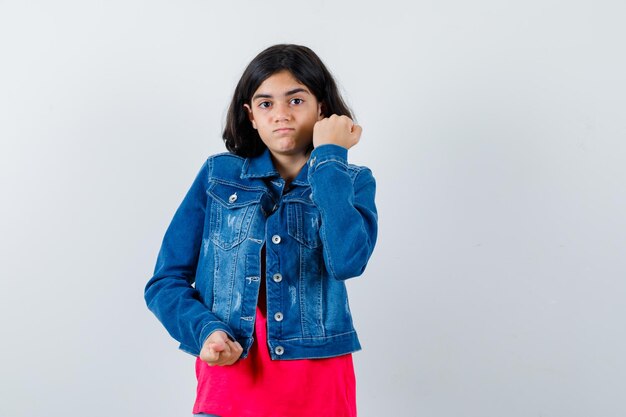 Young girl in red t-shirt and jean jacket clenching fist and looking serious , front view.