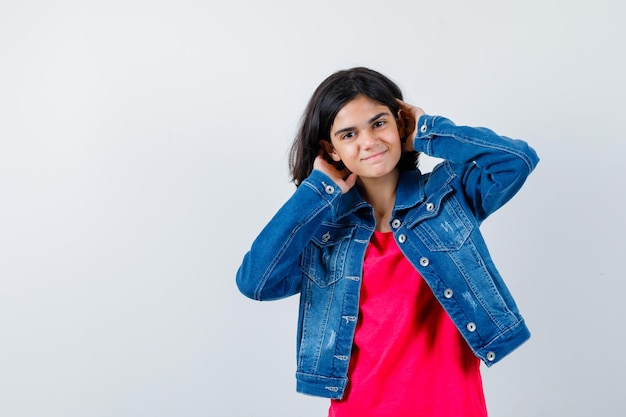 Young girl in red t-shirt and jean jacket holding hands behind head and looking happy