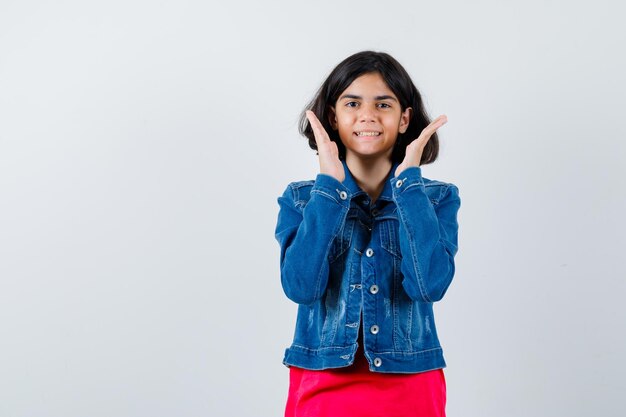 Young girl in red t-shirt and jean jacket holding hands near face and looking cheerful , front view.