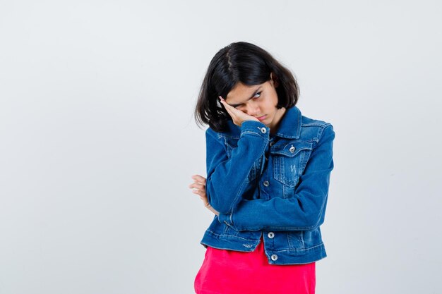 Young girl in red t-shirt and jean jacket leaning cheek on palm, thinking about something and looking pensive , front view.