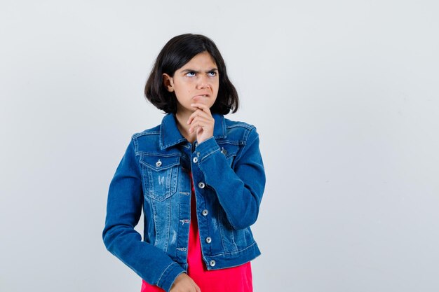 Young girl in red t-shirt and jean jacket leaning chin on palm while thinking about something and looking pensive , front view.