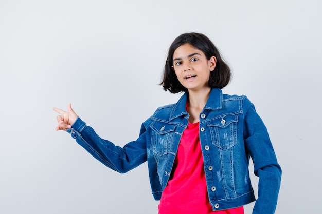 Young girl in red t-shirt and jean jacket pointing left with index finger and looking happy