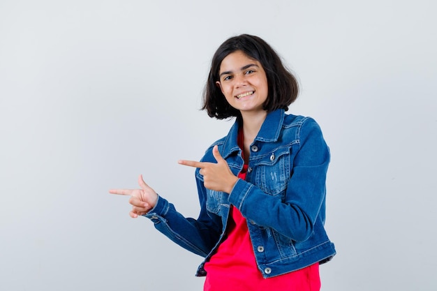 Young girl in red t-shirt and jean jacket pointing left with index fingers and looking happy , front view.