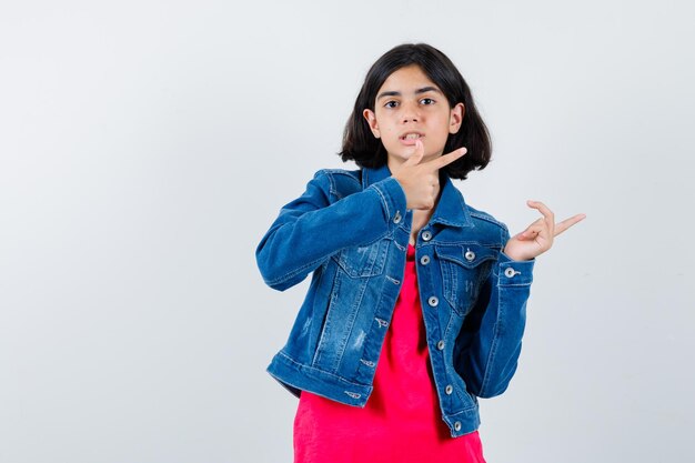 Young girl in red t-shirt and jean jacket pointing right with index fingers and looking cute , front view.