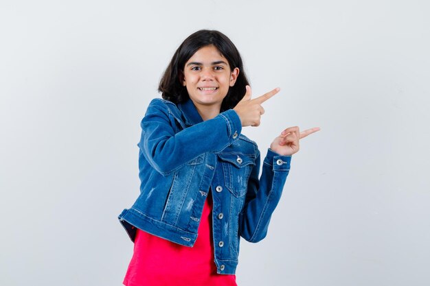 Young girl in red t-shirt and jean jacket pointing right with index fingers and looking happy , front view.