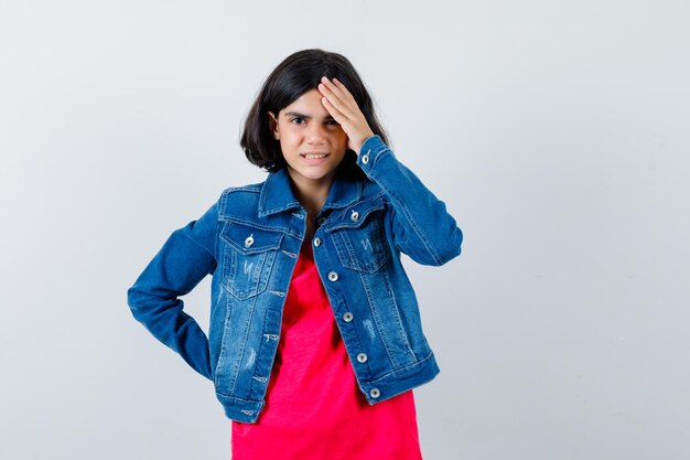 Young girl in red t-shirt and jean jacket putting hand on forehead while holding hand on waist and looking annoyed , front view.