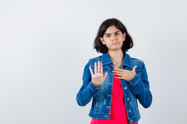 Giovane ragazza in maglietta rossa e giacca di jeans che mostra il segnale di stop e sembra seria, vista frontale.