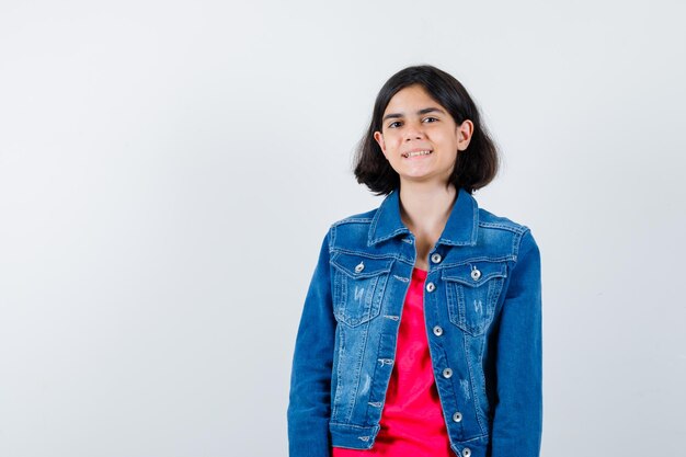 Young girl in red t-shirt and jean jacket standing straight and posing at camera and looking happy , front view.