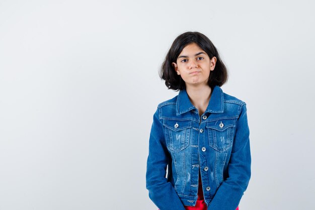 Young girl in red t-shirt and jean jacket standing straight and posing at camera and looking serious , front view.