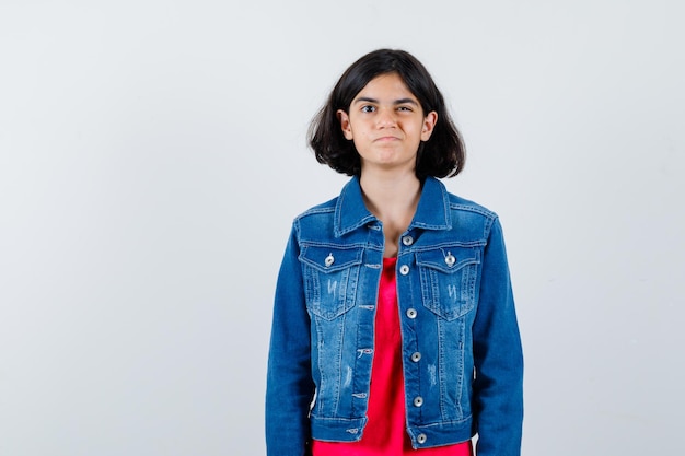 Giovane ragazza in maglietta rossa e giacca di jeans in piedi dritta, strizzando l'occhio e posando alla telecamera e sembrando carina