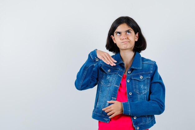 Young girl in red t-shirt and jean jacket standing in thinking pose and looking pensive , front view.