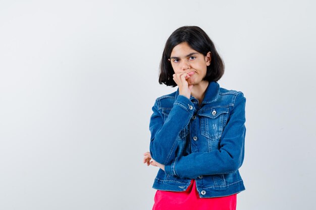 Young girl in red t-shirt and jean jacket standing in thinking pose and looking pensive , front view.