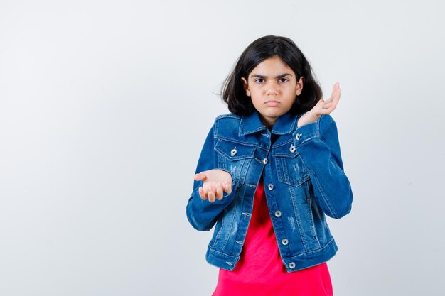 Young girl in red t-shirt and jean jacket stretching hands in questioning manner and looking baffled , front view.