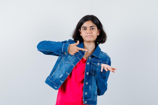 Young girl in red t-shirt and jean jacket stretching one hand as holding something and pointing to it with index finger and looking happy , front view.